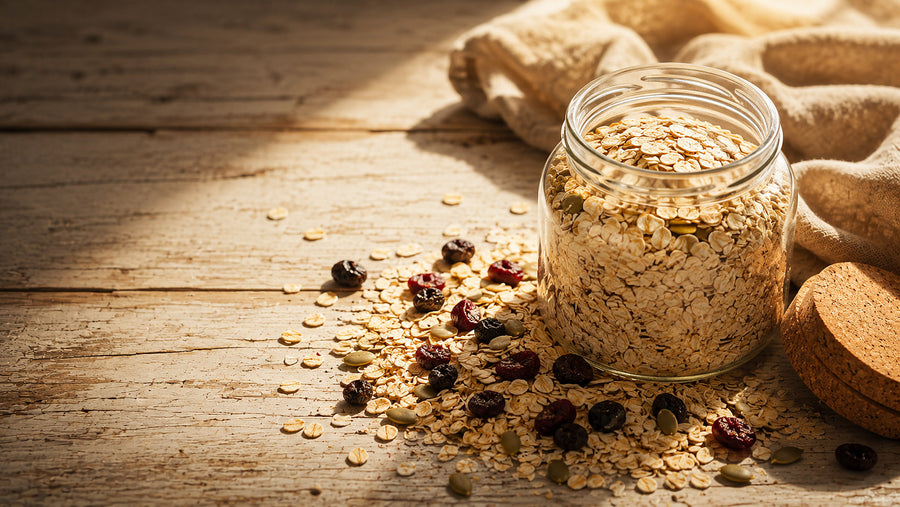 Open glass jar of rolled oats with a cork lid, dried berries and pumpkin seeds scattered across a pale wooden surface