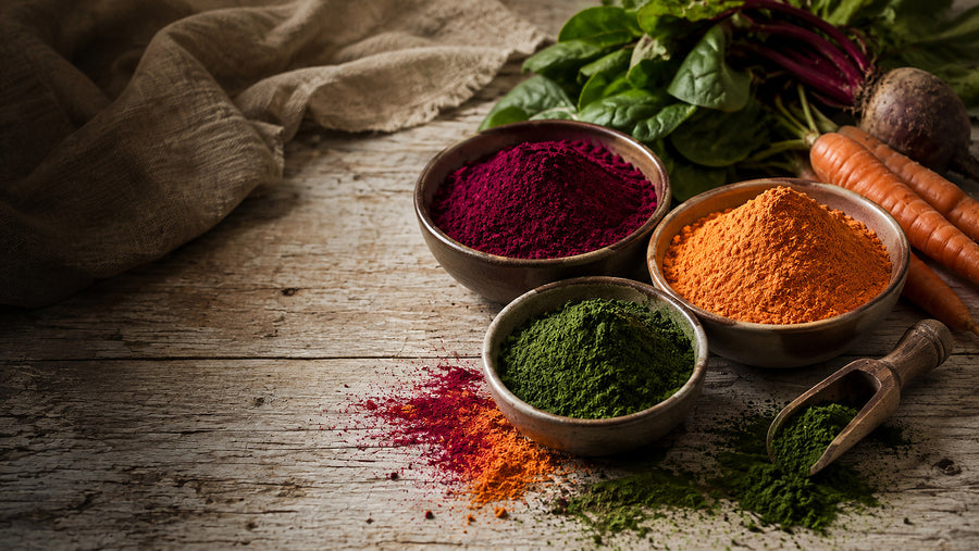 Three bowls of beetroot, carrot and spinach powder beside fresh beetroot, carrots and spinach on a pale wooden surface